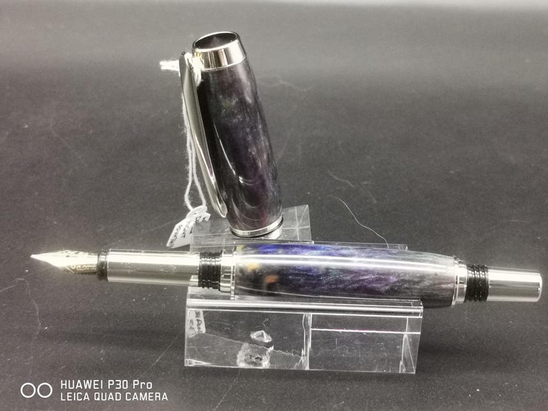 May include: A fountain pen with a silver nib and accents. The pen's body features a swirling pattern of purple, blue, and gray. The cap is open, revealing the nib. The pen is displayed on a clear acrylic stand.