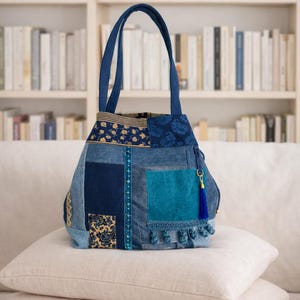 Puede incluir: Un bolso tote de mezclilla patchwork con una variedad de tonos azules, incluyendo índigo y verde azulado. El bolso presenta una borla azul y un ribete decorativo. El bolso está sobre dos almohadas beige.