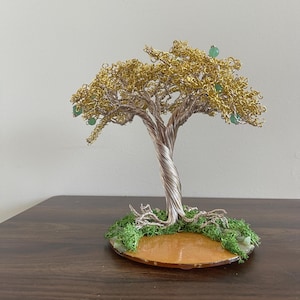 May include: A wire sculpture of a tree with a gold and silver finish. The tree has green gemstone leaves and is set on a wooden base with green moss.