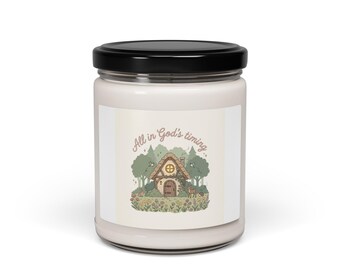 May include: A white candle jar with a black lid. The label features a whimsical illustration of a cottage with the text "All in God's timing." The cottage is surrounded by trees, flowers, and a deer. The candle is a home decor item.
