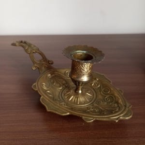 May include: An antique brass candlestick holder with an ornate, oval base and a handle. The candlestick features detailed floral and scrollwork patterns. The holder is designed to hold a single candle and is made of brass.