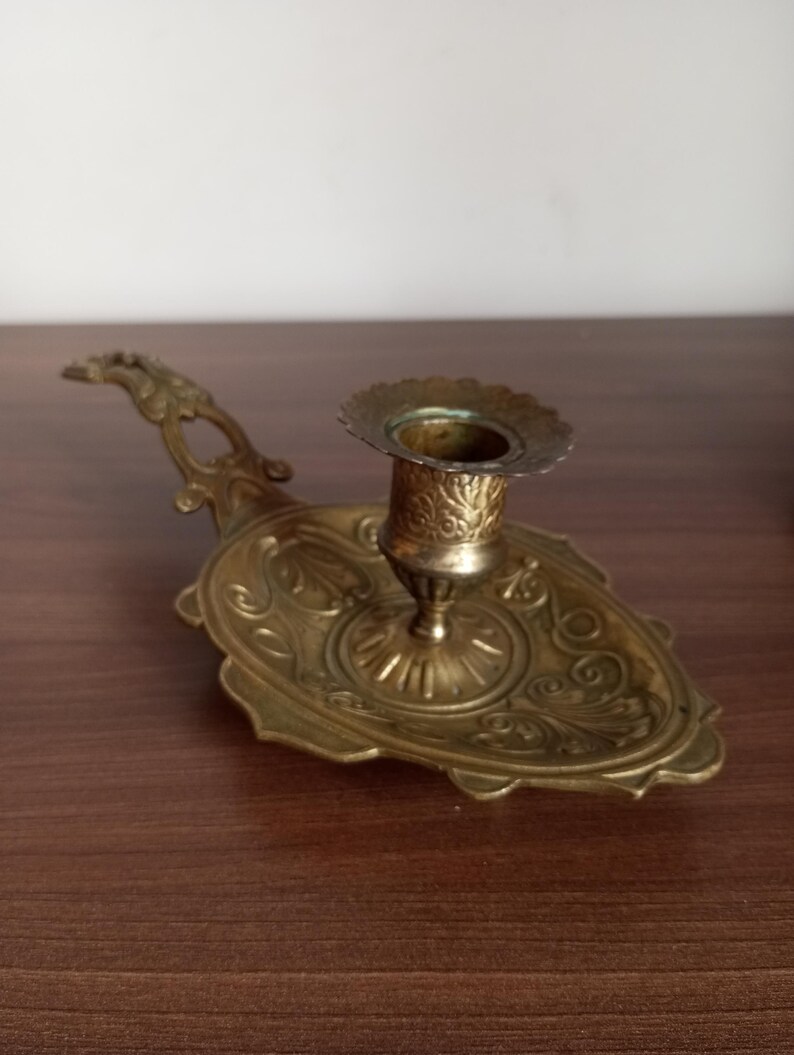 May include: An antique brass candlestick holder with an ornate, oval base and a handle. The candlestick features detailed floral and scrollwork patterns. The holder is designed to hold a single candle and is made of brass.
