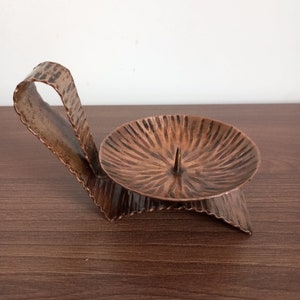May include: A hammered copper candle holder with a single candle holder and a curved handle. The holder has a textured surface and a rustic look.