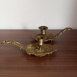 May include: Two antique brass candle holders with ornate detailing. Each holder features a circular base with a handle and a central cup for holding a candle. The brass has a warm, aged patina, suggesting vintage craftsmanship. The candle holders are displayed on a dark wooden surface.