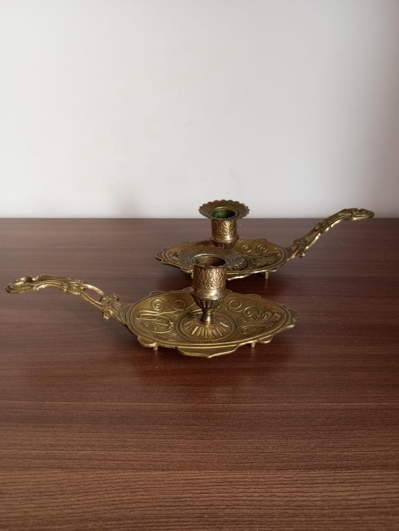 May include: Two antique brass candle holders with ornate detailing. Each holder features a circular base with a handle and a central cup for holding a candle. The brass has a warm, aged patina, suggesting vintage craftsmanship. The candle holders are displayed on a dark wooden surface.
