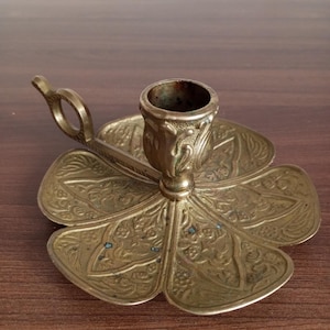 May include: A brass candlestick with a decorative floral design. The candlestick has a removable handle and a round base.