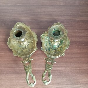 May include: Two antique brass candle holders. Each holder features an oval base with ornate floral detailing and a cup for the candle. The handles are also intricately designed. The candle holders are displayed on a wooden surface.