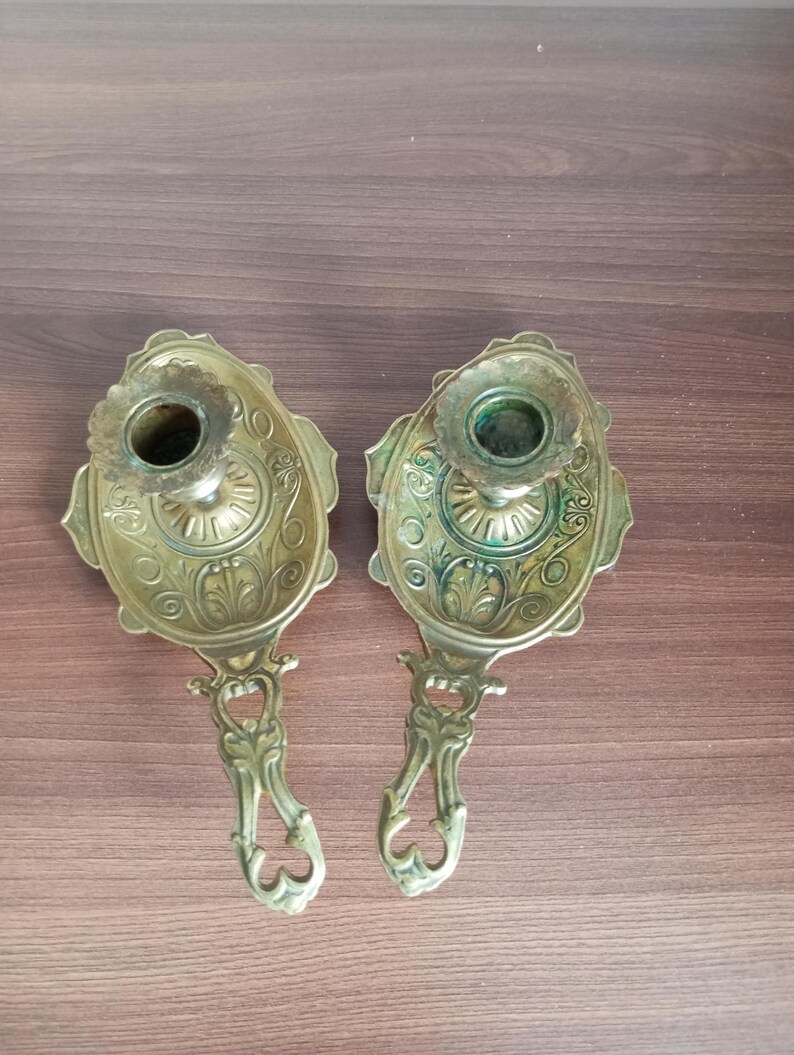 May include: Two antique brass candle holders. Each holder features an oval base with ornate floral detailing and a cup for the candle. The handles are also intricately designed. The candle holders are displayed on a wooden surface.