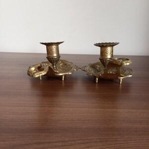 May include: Two antique brass candle holders. Each holder features a detailed floral design on the base and cup. The candle holders are connected by a decorative element. The brass has a warm, aged patina.