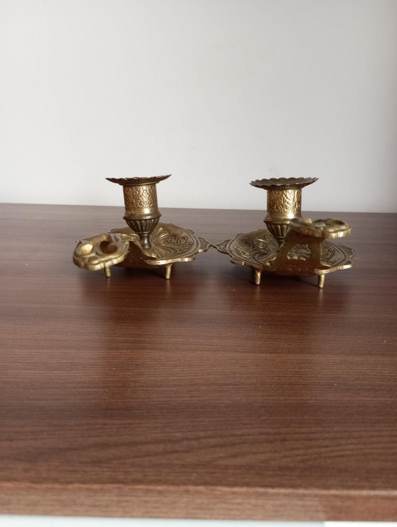 May include: Two antique brass candle holders. Each holder features a detailed floral design on the base and cup. The candle holders are connected by a decorative element. The brass has a warm, aged patina.