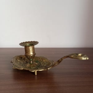 May include: Ornate brass candlestick holder with a decorative oval base and a handle. The candlestick features detailed floral and scrollwork patterns. The holder is designed to hold a single candle and has a small, scalloped rim.