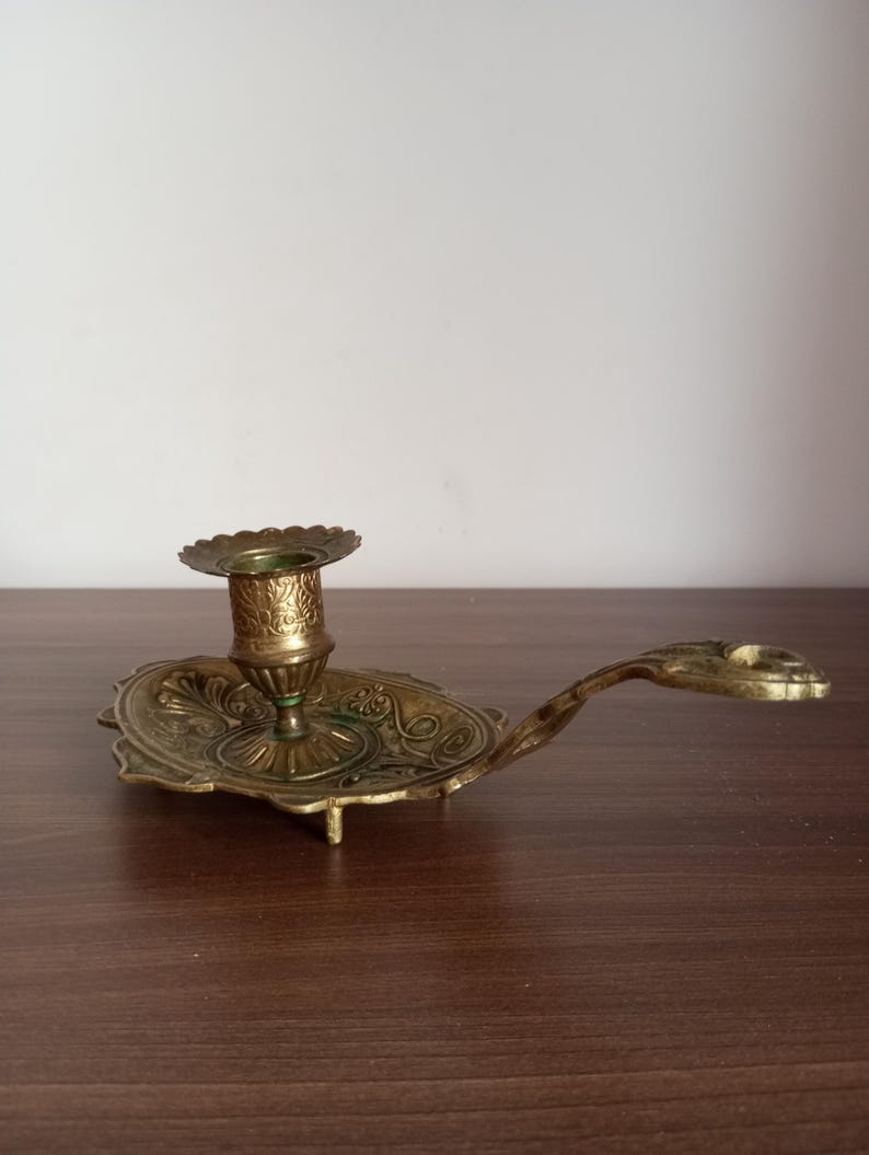 May include: Ornate brass candlestick holder with a decorative oval base and a handle. The candlestick features detailed floral and scrollwork patterns. The holder is designed to hold a single candle and has a small, scalloped rim.