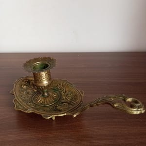 May include: Antique brass candlestick holder with an ornate design. The candle holder features a circular base with an oval tray and a handle. The design includes floral and scrollwork patterns, and the brass has a slightly aged patina.