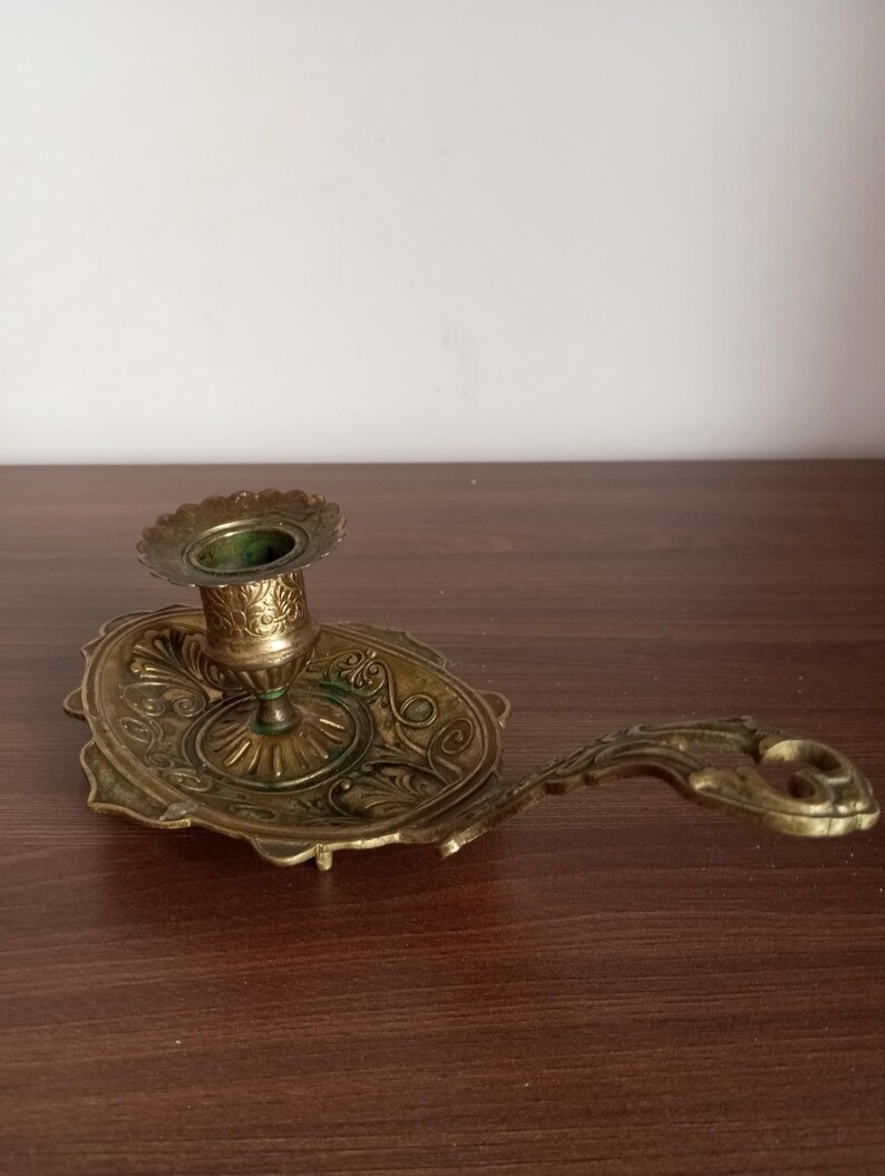 May include: Antique brass candlestick holder with an ornate design. The candle holder features a circular base with an oval tray and a handle. The design includes floral and scrollwork patterns, and the brass has a slightly aged patina.