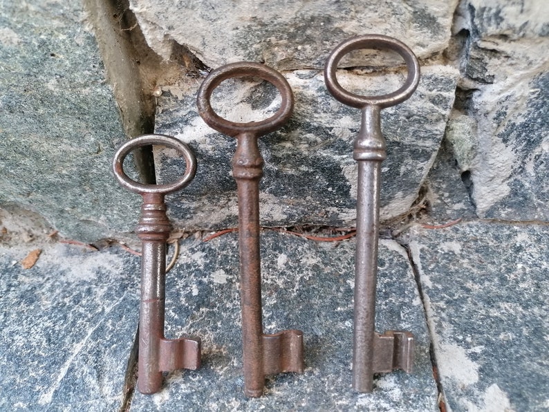 K&ouml;nnte beinhalten: Drei antike Schl&uuml;ssel mit verziertem Metall. Die Schl&uuml;ssel sind aus dunklem Metall und haben ein rustikales Aussehen. Sie sind vertikal vor einem Stein Hintergrund angeordnet.