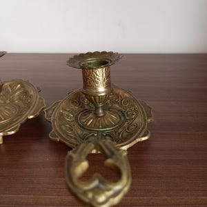 May include: Antique brass candle holders with ornate floral designs. The candle holders have a scalloped edge and a handle. The brass has a vintage patina. The candle holders are on a dark brown surface.