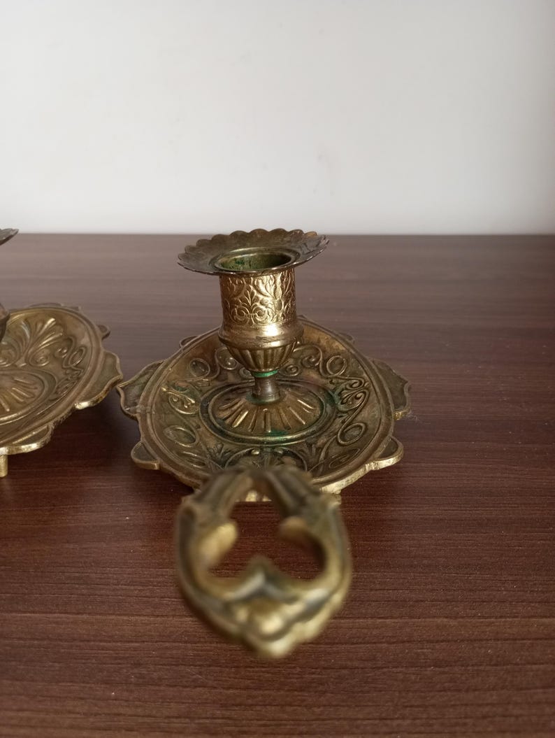May include: Antique brass candle holders with ornate floral designs. The candle holders have a scalloped edge and a handle. The brass has a vintage patina. The candle holders are on a dark brown surface.