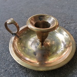 May include: A brass candlestick with a round base and a single candle holder. The candlestick has a handle on the side.