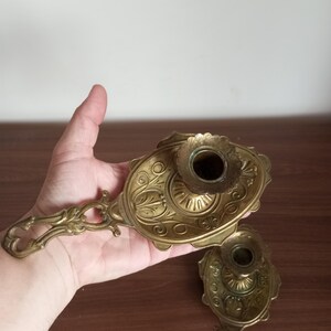 May include: Two antique brass candle holders with ornate floral and scrollwork designs. Each holder features a handle and a circular cup for holding a candle. The brass has a warm, aged patina, suggesting vintage craftsmanship.