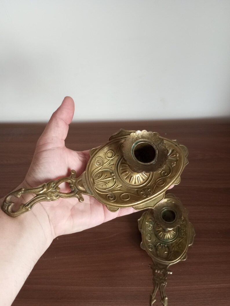 May include: Two antique brass candle holders with ornate floral and scrollwork designs. Each holder features a handle and a circular cup for holding a candle. The brass has a warm, aged patina, suggesting vintage craftsmanship.