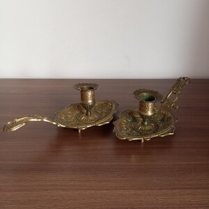 May include: Two antique brass candle holders. Each holder features an ornate, oval base with a detailed floral design and a small cup to hold a candle. The holders have a long, curved handle, and are displayed on a wooden surface.