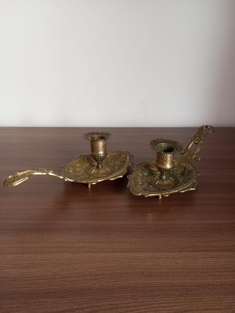 May include: Two antique brass candle holders. Each holder features an ornate, oval base with a detailed floral design and a small cup to hold a candle. The holders have a long, curved handle, and are displayed on a wooden surface.