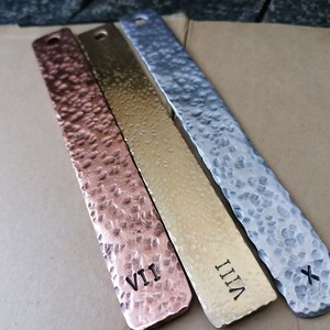 Puede incluir: Tres marcapáginas de metal con textura martillada. Los marcapáginas están etiquetados con los números romanos VII, VIII y X. Los marcapáginas son de color cobre, oro y plata.