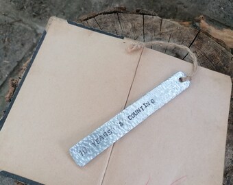 Könnte beinhalten: Ein silbernes Metall-Lesezeichen mit dem eingravierten Text "10 YEARS & COUNTING". Das Lesezeichen ist mit einer braunen Schnur gebunden.
