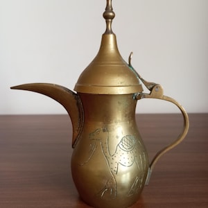 Puede incluir: Una cafetera de latón vintage con un pico largo, un asa curvada y un grabado decorativo de camello. La cafetera tiene una tapa cónica con un remate puntiagudo. El latón tiene una pátina cálida y envejecida.