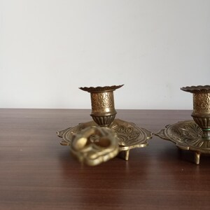 May include: Ornate brass candle holders with detailed floral patterns. The candle holders have a round base with a scalloped edge and a central cup to hold the candle. The brass has a warm, antique finish.