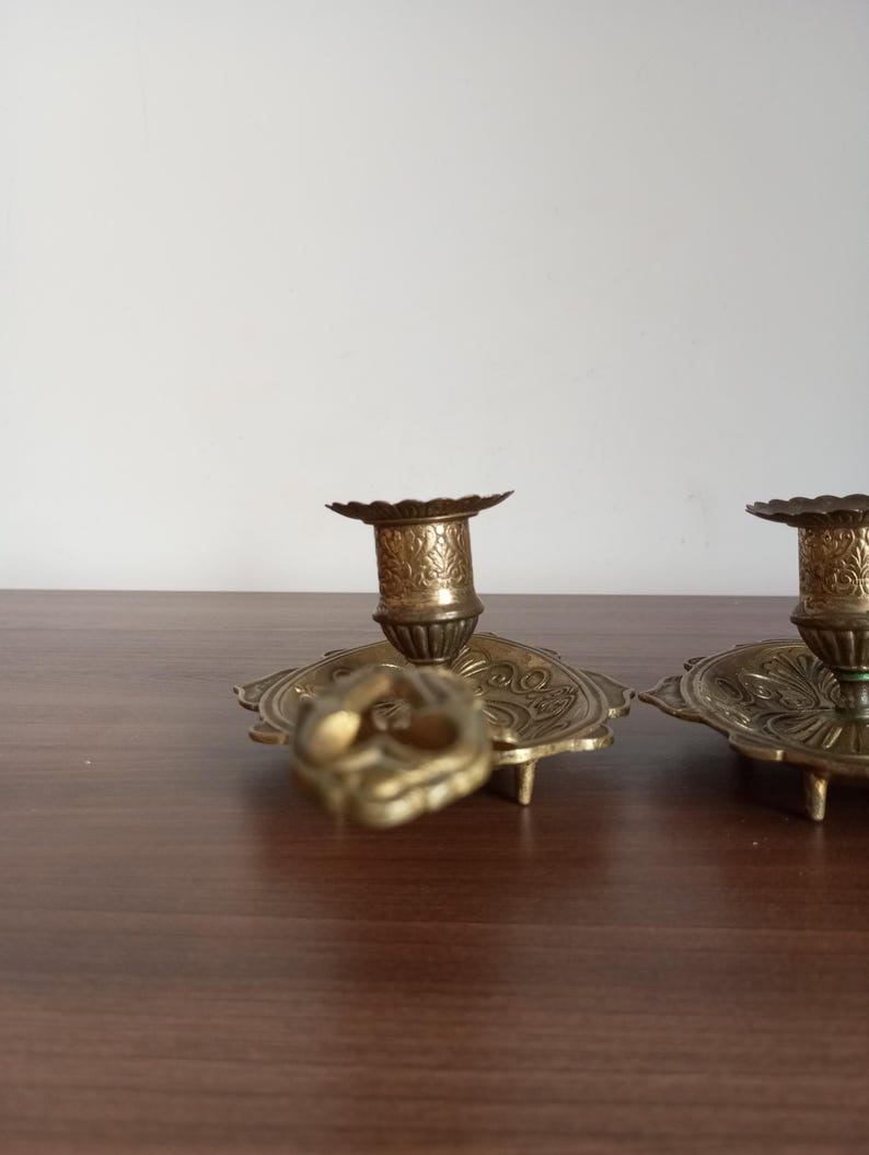 May include: Ornate brass candle holders with detailed floral patterns. The candle holders have a round base with a scalloped edge and a central cup to hold the candle. The brass has a warm, antique finish.