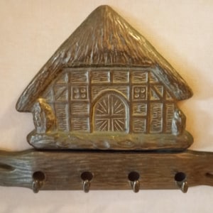 Puede incluir: Un llavero rústico con forma de cabaña con tejado de paja. El llavero está hecho de un material color bronce y tiene cuatro ganchos debajo de la cabaña. La cabaña tiene un diseño detallado de puerta y ventana.