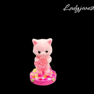 May include: Pink plush cat figurine holding a pink lollipop, standing on a pink and white base with colorful confetti.