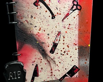 Könnte beinhalten: Ein pinkfarbenes und schwarzes Journal mit weißem Hintergrund und einem roten Spritzmuster. Das Journal hat eine schwarze Ringbindung und einen schwarzen Plastikgrabstein mit dem Wort "RIP" darauf. Das Journal ist mit schwarzen Silhouetten eines Messers, einer Axt und einer Schere verziert.