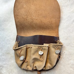 May include: A tan leather pouch with a dark brown leather trim and flap. The pouch has three small pockets with decorative closures. The flap is open, revealing the interior. The pouch is designed to be worn on a belt.