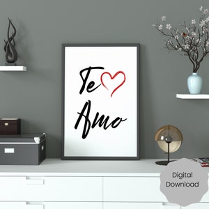 Op de afbeelding: Een printbare wandkunstprint met de tekst "Te Amo" in zwarte schrijfletters met een rood hart. De print is ingelijst in een zwart frame en wordt getoond op een wit rek.