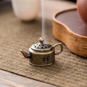 May include: A small, antique-style brass teapot incense burner with the Chinese character for "Zen" on the front. Smoke rises from the top. The burner sits on a textured, brown surface.