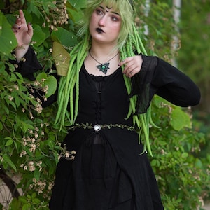 May include: A person with green hair and a black dress is standing in front of a green leafy background. They are wearing a green gemstone necklace and a belt with a silver buckle.