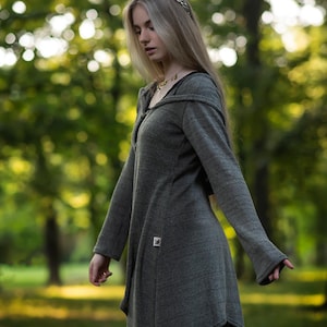 Puede incluir: Vestido gris con capucha y mangas largas, con un dobladillo asimétrico. El vestido tiene cierre de cremallera y un pequeño parche cuadrado. La modelo lleva leggings negros y una diadema plateada, posando al aire libre.