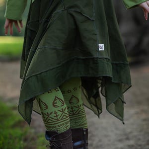 Puede incluir: Falda verde oliva oscuro con paneles asimétricos en capas. La falda se combina con leggings estampados en tonos verdes y marrones, y botas marrones oscuras con cordones. El conjunto tiene un tema bohemio o de fantasía.