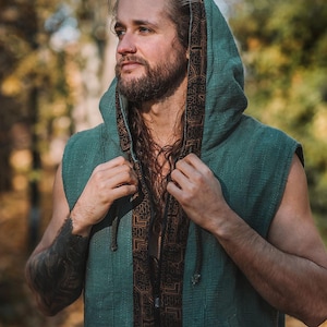 May include: A sleeveless green hooded vest with a brown patterned trim. The vest has a zipper closure and a drawstring hood. The person in the image is wearing the vest outdoors.