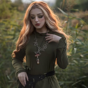 May include: A woman with long, wavy, blonde hair wears a green, long-sleeved shirt with a black belt and a black leather fanny pack. She is wearing a necklace with a mushroom pendant. The background is a field of tall grass.