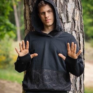 Puede incluir: Sudadera con capucha negra con un diseño estampado en la capucha y la parte inferior delantera. La sudadera se combina con pantalones verdes con cordón. La persona de la imagen está de pie junto a un árbol.