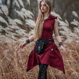 Puede incluir: Vestido con capucha burdeos con cinturón y bolsa negros, combinado con leggings negros y botas marrones. El vestido sin mangas tiene un dobladillo asimétrico. El atuendo se complementa con pulseras de muñeca. La modelo está de pie en un campo de hierba alta.