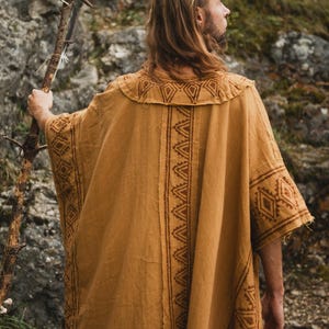 Op de afbeelding: Een persoon draagt een mosterdkleurige poncho met geometrische patronen en een decoratieve staf. De poncho heeft ingewikkelde bruine ontwerpen langs de randen en op de rug. De staf is gemaakt van hout en heeft een uniek, natuurlijk ontwerp.