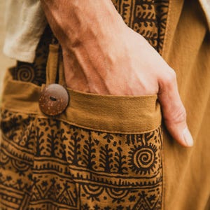 Può includere: Primo piano di pantaloni color cachi con un motivo marrone. I pantaloni hanno un grande bottone di legno e una tasca. Il design include forme di alberi e sole. Una mano è in tasca.