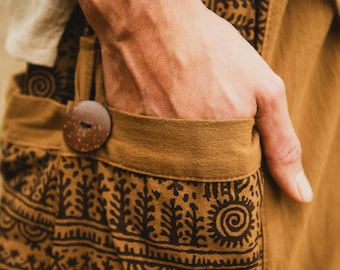 May include: Close-up of tan pants with a brown, patterned design. The pants have a large wooden button and a pocket. The design includes tree and sun-like shapes. The person's hand is in the pocket.