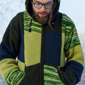 Könnte beinhalten: Ein Kapuzenpullover mit Reißverschluss in einem Patchwork-Design. Der Pullover hat eine Mischung aus grünen, marineblauen und schwarzen Paneelen. Die Kapuze ist hochgezogen, und die Person trägt eine Brille. Der Pullover hat einen Kordelzug und zwei Vordertaschen.