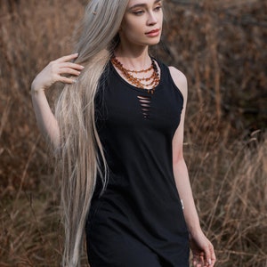 Peut inclure: Une femme aux longs cheveux blonds portant une robe noire sans manches avec un décolleté en détresse. Elle porte un collier superposé de perles d'ambre.