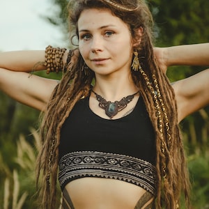 May include: A black crop top featuring a wide band with a geometric pattern, paired with a macrame necklace containing a central dark stone. The jewelry includes dangling hand-shaped earrings and a beaded bracelet on the wrist.